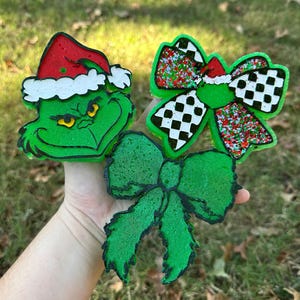 May include: Three Christmas-themed decorations. One is a green Grinch face wearing a red Santa hat. The other two are green bows, one with a black and white checkered pattern and the other a solid green. All have a glittery texture.