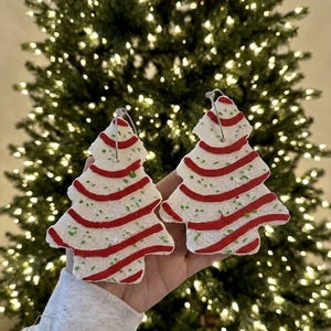 May include: Two white Christmas tree-shaped ornaments with textured surfaces, red horizontal stripes, and small green speckles. Each ornament features a silver metal loop at the top for hanging, designed to resemble frosted holiday cookies.