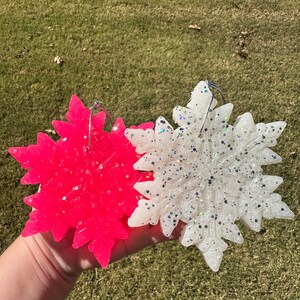 May include: Two snowflake ornaments, one bright pink and one white, are held against a green background. Both ornaments are covered in glitter and have silver wire hangers. The snowflake design is detailed and intricate.
