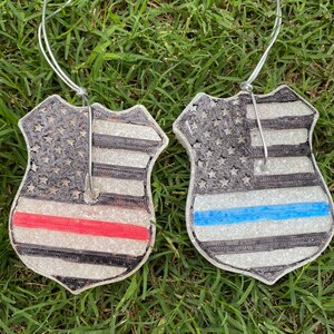 May include: Two silver car air fresheners shaped like police badges with an American flag design. One has a red stripe across the flag and the other has a blue stripe.
