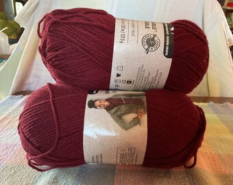 May include: Two burgundy yarn skeins are stacked on top of each other. Each skein has a label with text and a picture of a person wearing a scarf. The yarn is Loops & Threads brand, and each skein is 253 m / 127.5 g.