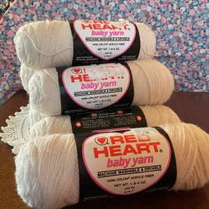 May include: Four skeins of Red Heart baby yarn in a soft, off-white color. Each skein is labeled with the brand name, fiber content, and net weight of 1.75 oz. The yarn is machine washable and dryable.