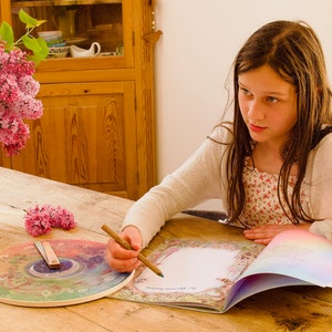 May include: A young person is seated at a wooden table, writing in a journal. A yellow pitcher with pink flowers sits to the left. A decorative clock and open book are on the table. The journal has a floral border and the text "My Morning Routine".