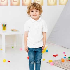 May include: A young boy with curly blonde hair is standing in a room with white walls and a light grey rug. He is wearing a white polo shirt and blue jeans. There are colourful blocks and balls scattered on the floor. The letters D, E, F, G, H, and I are displayed on the wall behind him.