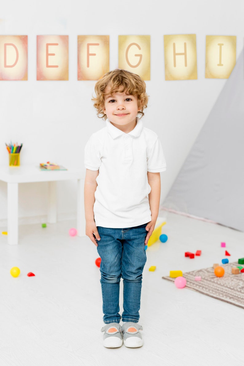 May include: A young boy with curly blonde hair is standing in a room with white walls and a light grey rug. He is wearing a white polo shirt and blue jeans. There are colourful blocks and balls scattered on the floor. The letters D, E, F, G, H, and I are displayed on the wall behind him.