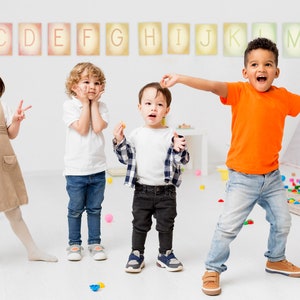 May include: Four children, two girls and two boys, are playing in a classroom. The children are standing in front of a white wall with colourful letters of the alphabet displayed on it. The children are wearing casual clothing and are smiling and laughing.