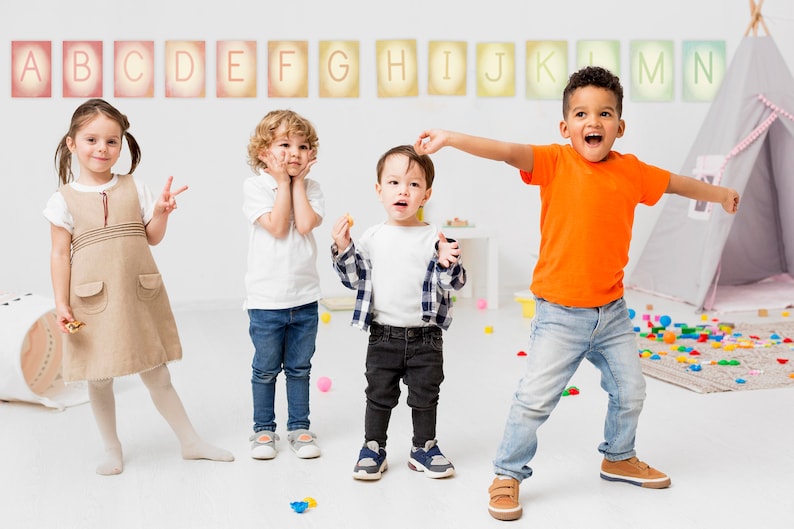 May include: Four children, two girls and two boys, are playing in a classroom. The children are standing in front of a white wall with colourful letters of the alphabet displayed on it. The children are wearing casual clothing and are smiling and laughing.