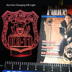 May include: A red, color-changing LED light shaped like a New York City Police badge with the text "K9-11". A magazine titled "Police K9" is visible in the background. The light is approximately 6 inches tall.