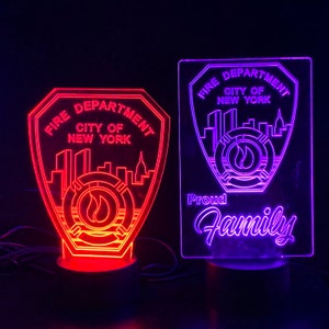 May include: Two illuminated LED signs with the New York City Fire Department logo. The sign on the left is red and the sign on the right is purple. The purple sign also has the text "Proud Family" below the logo.