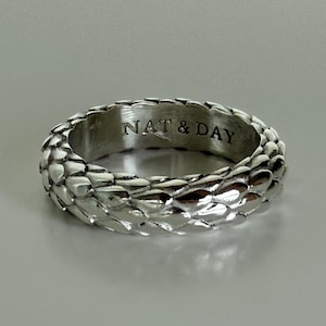May include: A silver ring with a textured, scale-like pattern. The ring is engraved with the text "NAT & DAY" on the inside. The ring is a band style and appears to be made of metal.