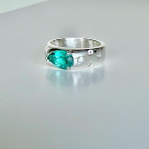 May include: A silver ring featuring a prominent pear-shaped teal gemstone set into the band. The band is accented with four small, round, clear stones embedded into the metal surface, arranged in a cluster near the primary teal gemstone.