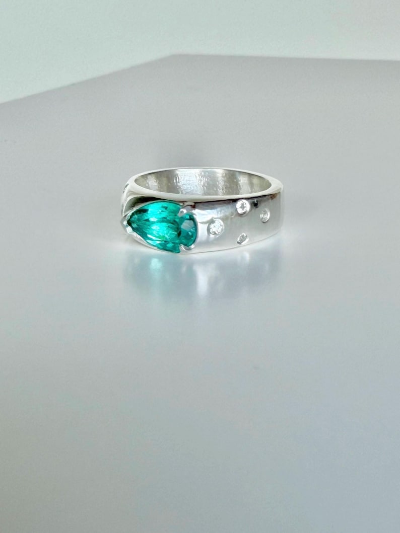 May include: A silver ring featuring a prominent pear-shaped teal gemstone set into the band. The band is accented with four small, round, clear stones embedded into the metal surface, arranged in a cluster near the primary teal gemstone.