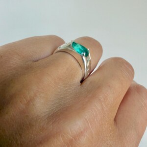 May include: A silver ring featuring a central marquise-cut vibrant green gemstone set in a polished metal band. The band is accented with small, circular embedded stones along the sides, creating a sleek and reflective appearance on the finger.