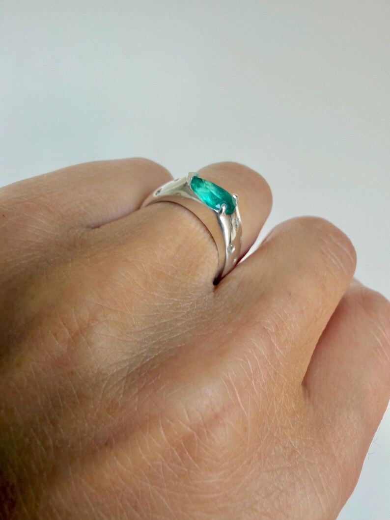 May include: A silver ring featuring a central marquise-cut vibrant green gemstone set in a polished metal band. The band is accented with small, circular embedded stones along the sides, creating a sleek and reflective appearance on the finger.
