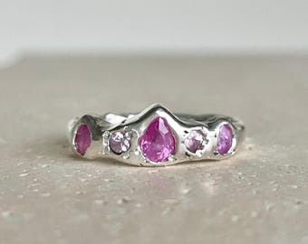 Puede incluir: Un anillo de plata con diseño de corona, adornado con piedras preciosas rosas. El anillo tiene una banda texturizada y está engastado con una piedra central en forma de pera, flanqueada por gemas rosas redondas y ovaladas más pequeñas. El anillo se muestra sobre una superficie neutra.