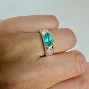 May include: A silver ring featuring a pear-shaped, bright teal gemstone set in a smooth, polished band. The band includes small, circular indentations or accents surrounding the central stone, creating a textured appearance on the metal surface.
