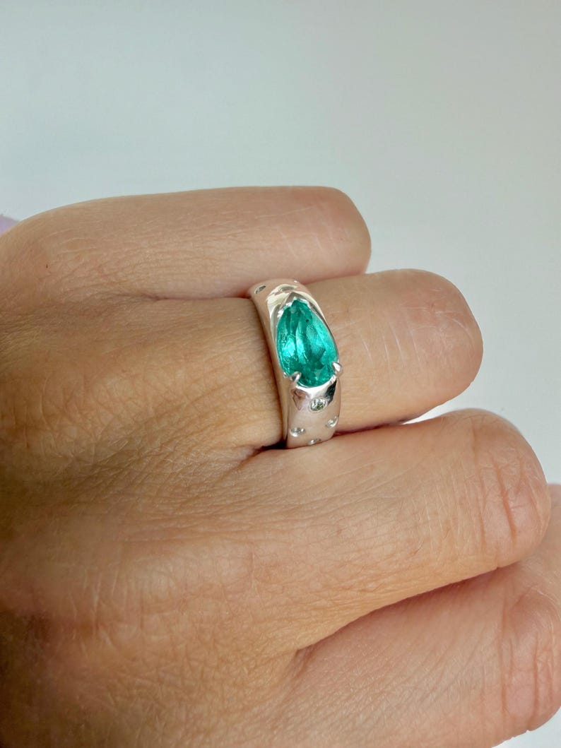 May include: A silver ring featuring a pear-shaped, bright teal gemstone set in a smooth, polished band. The band includes small, circular indentations or accents surrounding the central stone, creating a textured appearance on the metal surface.