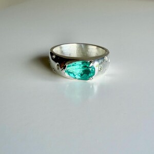 May include: A polished silver band ring featuring a central pear-shaped, faceted teal-green gemstone held by prongs. The wide, rounded metal band is embedded with several small, clear, circular stones set flush into the silver surface around the center.