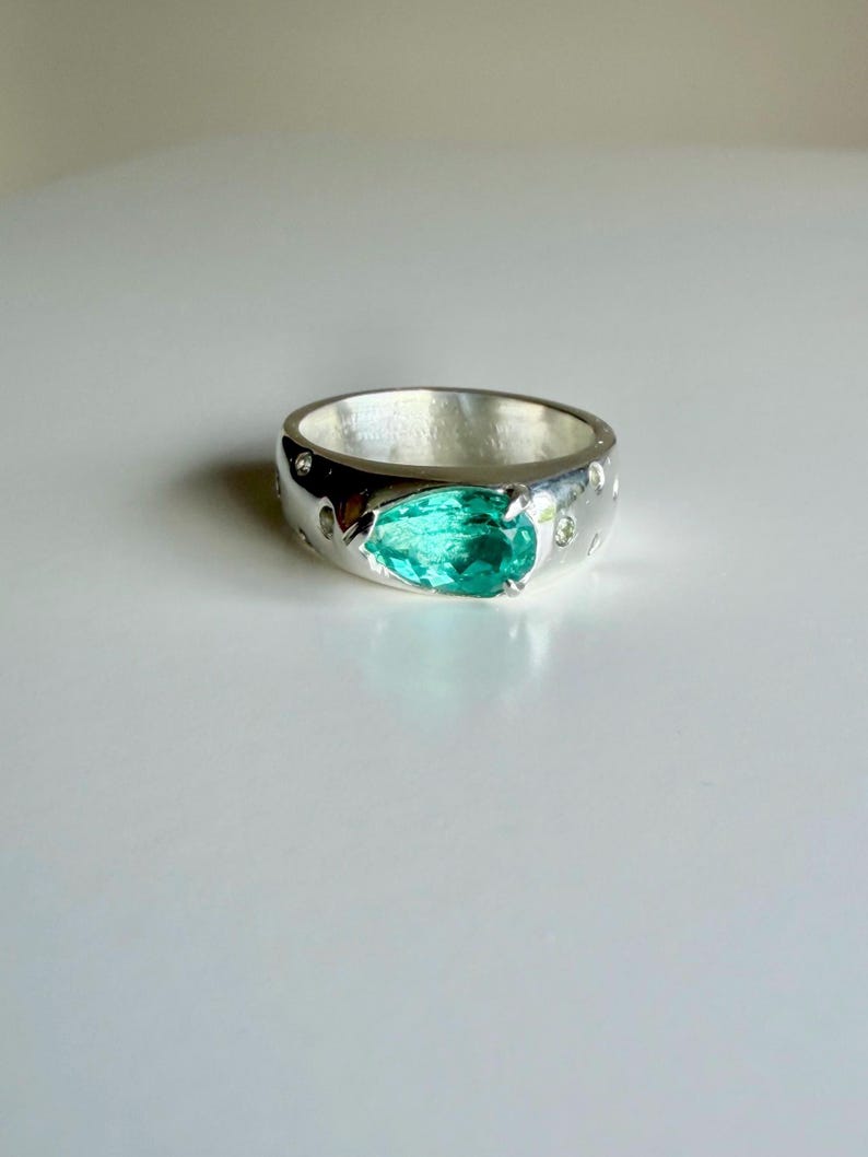 May include: A polished silver band ring featuring a central pear-shaped, faceted teal-green gemstone held by prongs. The wide, rounded metal band is embedded with several small, clear, circular stones set flush into the silver surface around the center.