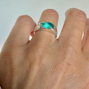 May include: A silver ring featuring a prominent, pear-shaped, vibrant green gemstone set in the center. The band has a smooth, polished finish and is accented with several small, round, clear stones embedded along the sides of the metal band.