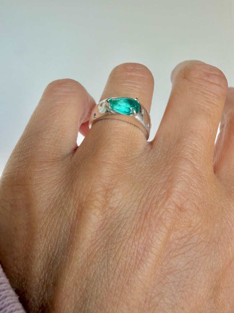 May include: A silver ring featuring a prominent, pear-shaped, vibrant green gemstone set in the center. The band has a smooth, polished finish and is accented with several small, round, clear stones embedded along the sides of the metal band.