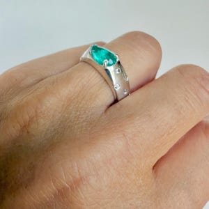 May include: A silver-toned ring featuring a pear-shaped green gemstone set at the top, accented by several small, round, clear stones embedded along the band. The band has a smooth, polished finish and a wide, solid construction.