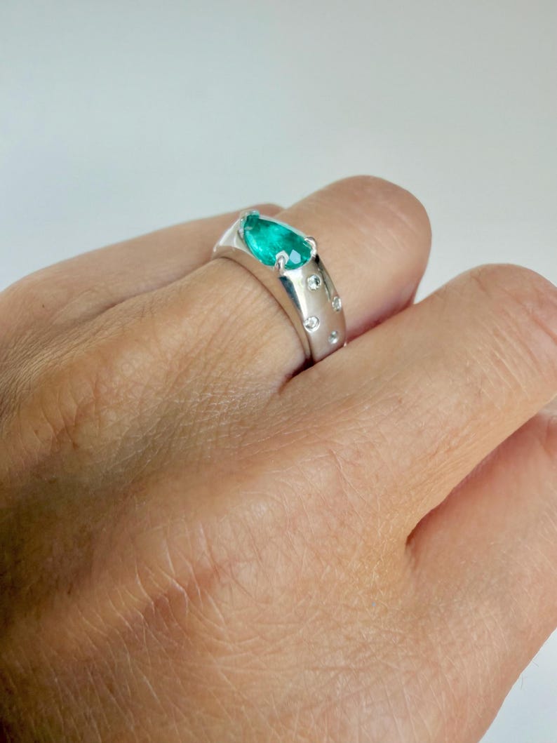 May include: A silver-toned ring featuring a pear-shaped green gemstone set at the top, accented by several small, round, clear stones embedded along the band. The band has a smooth, polished finish and a wide, solid construction.