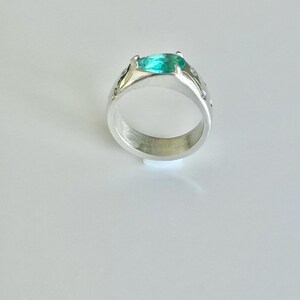 May include: A silver ring featuring a pear-shaped, bright teal gemstone set in a prong setting. The band is wide and polished, accented with small, clear, round stones embedded along the sides of the metal shank, creating a reflective surface.