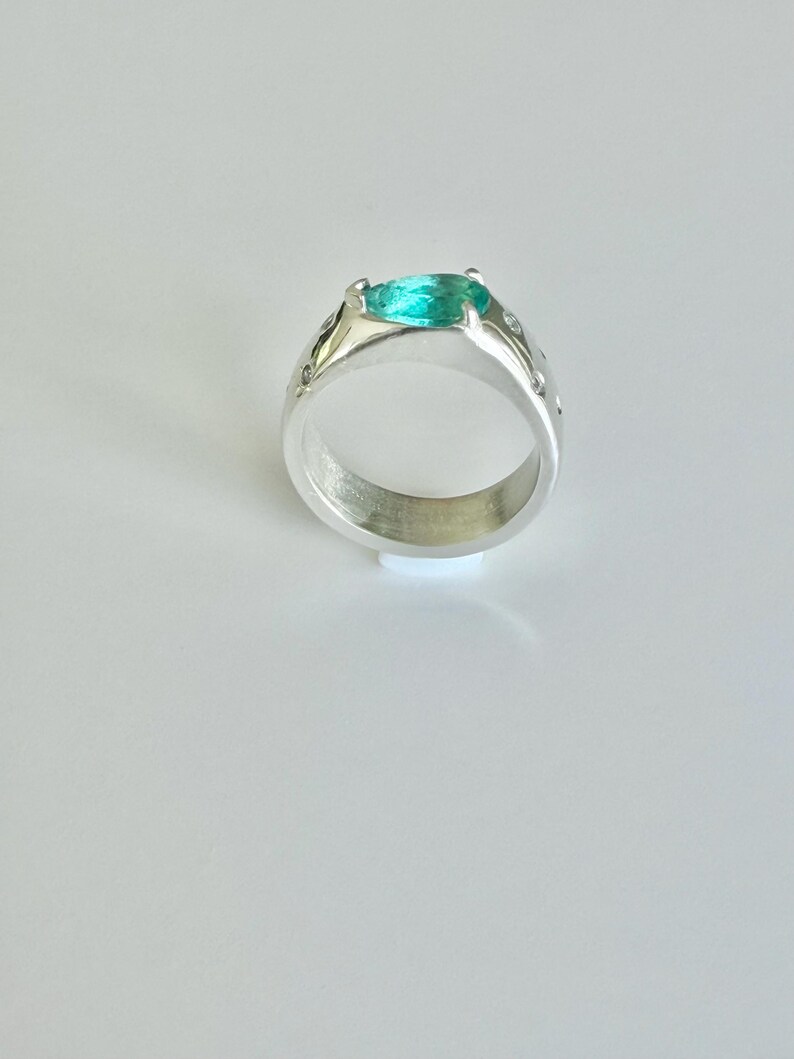 May include: A silver ring featuring a pear-shaped, bright teal gemstone set in a prong setting. The band is wide and polished, accented with small, clear, round stones embedded along the sides of the metal shank, creating a reflective surface.