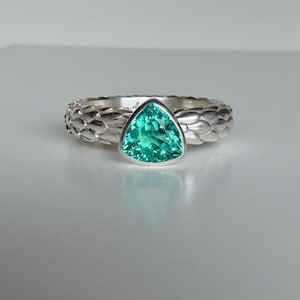 Puede incluir: Un anillo de plata con una gema triangular de color verde esmeralda. La banda tiene un diseño texturizado en forma de escamas. El anillo se encuentra sobre un fondo neutro, resaltando el color vibrante de la gema.
