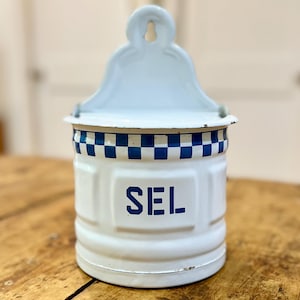 May include: A white enameled metal wall-mounted salt container featuring a blue and white checkered border near the top rim. The front of the cylindrical canister is embossed with the word SEL in bold blue capital letters.