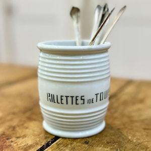 May include: A white ceramic crock with horizontal ribbed detailing and the black text RILLETTES DE TOURS printed on the front. The cylindrical container holds several silver-toned metal spoons standing upright inside the open top.