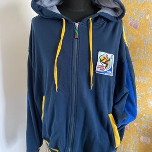 May include: Navy blue zip-up hoodie with a gray interior hood lining and yellow drawstrings. Features a patch with the South Africa 2010 FIFA World Cup logo. The sleeves have blue and yellow accents. A casual, sporty garment.