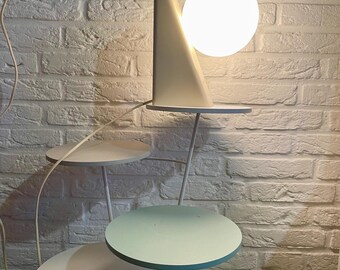 May include: A white lamp with a round, white shade sits on a white shelf. The lamp is attached to a white metal stand with three circular shelves. The shelves are painted white, light blue, and white.