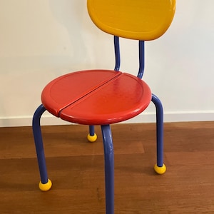 May include: A colorful children's chair with a round red seat and a yellow backrest. The chair has blue legs and yellow feet. The chair is made of metal and has a retro design.