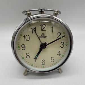 Könnte beinhalten: Ein silberner Wecker mit weißem Zifferblatt und schwarzen Zeigern. Die Uhr hat eine verchromte Oberfläche und ein klares Glaszifferblatt. Das Zifferblatt der Uhr hat schwarze Zahlen und einen grünen Punkt an jeder Stundenmarkierung. Die Uhr wird von Peter hergestellt.