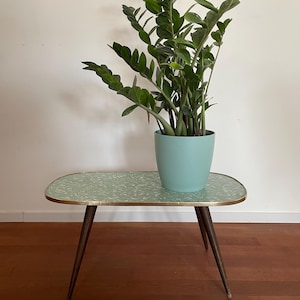 May include: A small, mid-century modern style side table with a light green patterned top and brass trim. The table has dark brown, tapered legs. A light blue pot with a green plant sits on top of the table.