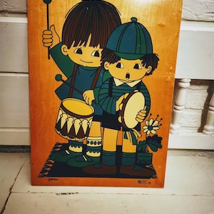 May include: A vintage wooden plaque with a colorful illustration of two children playing music. One child is playing a drum and the other is playing a tambourine. The plaque has a brown wood background and the children are drawn in a cartoon style with bright colors.