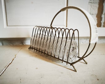 May include: A gold metal coaster rack with a white woven handle. The rack holds a set of clear glass coasters with a decorative pattern.