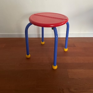 May include: A small, colorful stool with a red, round seat and blue legs. The legs are capped with yellow spheres. The stool is on a wooden floor and against a white wall.