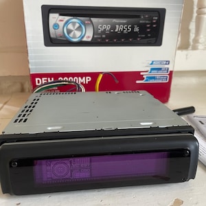 Könnte beinhalten: Ein Pioneer DEH-2000MP Autoradio mit schwarzer Frontblende und einem violetten digitalen Display. Das Gerät steht vor seiner originalen weiß-roten Kartonverpackung, die eine Abbildung des Stereo-Systems zeigt.