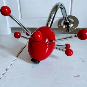 May include: A red and chrome wall-mounted coat rack. The rack has a red, egg-shaped body with chrome arms ending in red spheres. A curved chrome arm connects to a circular mounting plate.
