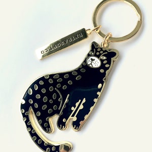 Può includere: Portachiavi dorato con un leopardo in smalto nero e oro con occhi bianchi. Il portachiavi include anche un'etichetta dorata con il testo "EXTRASOFT.CO".