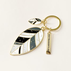 May include: A gold-tone keychain featuring a black, white and grey leaf design. The keychain also includes a smaller leaf charm and a rectangular tag with the text "EXTRASOFT.CO".