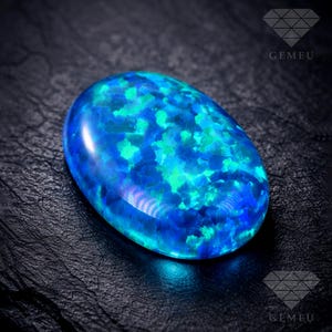 Può includere: Una gemma ovale con un vivace motivo di colore blu e verde. La superficie lucida riflette la luce, mostrando un design a mosaico. La gemma è posta su uno sfondo scuro e strutturato.