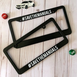 May include: Two black license plate frames with the text "#SAVETHEMANUALS" in white letters. The frames are on a light wood background.
