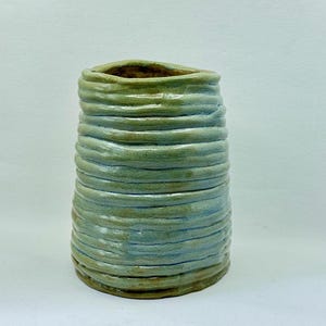 May include: A cylindrical ceramic vase with a hand-coiled, textured surface. The exterior features a glossy glaze in shades of sage green and light blue, with visible horizontal ridges created by the stacked clay coils and a wide, open top.