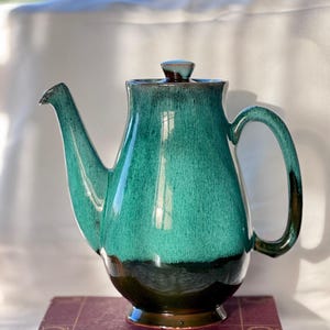 May include: A teal and brown ceramic teapot with a curved handle and spout. The teapot sits on a vintage book with the title "Holmes' Complete Poems". The teapot has a glossy finish and a lid with a small knob.