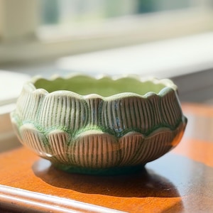 May include: A ceramic bowl featuring a sculpted lotus flower design with a scalloped rim. The exterior displays a light green glaze with a subtle crackle finish and vertical ribbed textures on each petal, while the interior is a smooth light green.