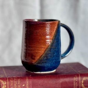 May include: A handmade ceramic mug with a unique color scheme. The mug features a diagonal split design with a brown and blue color palette. The mug is sitting on top of a vintage book with gold lettering.